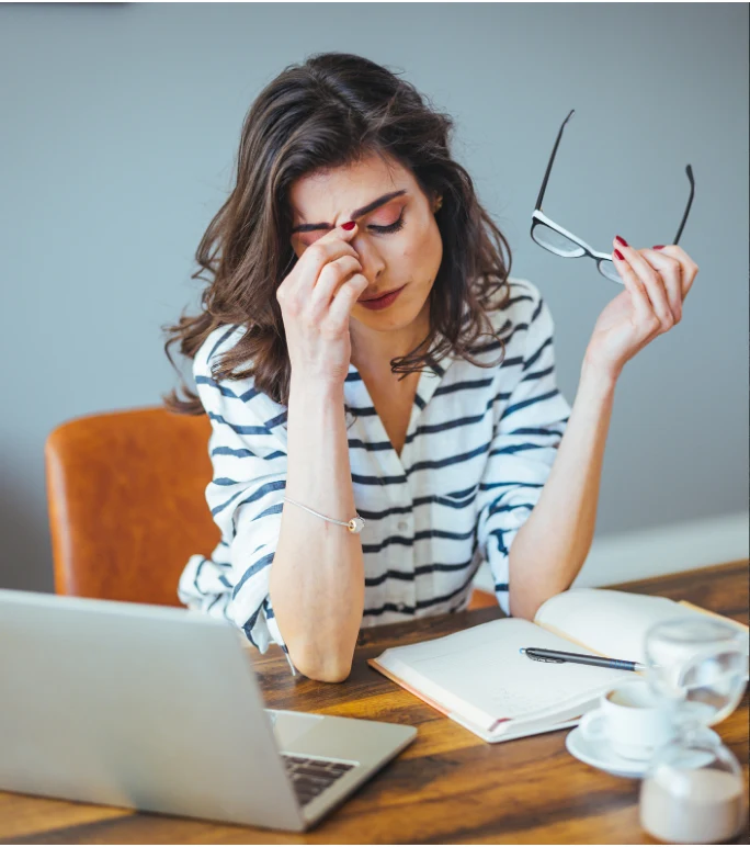 Teacher sitting at a desk with a laptop and notebook, holding her glasses and rubbing her eyes